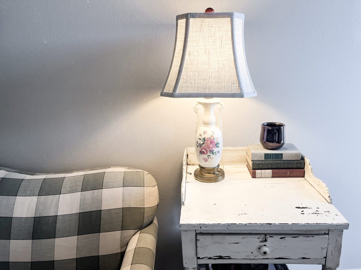 Floral vintage lamp next to stack of old books on a distressed white table next to green checkered vintage couch