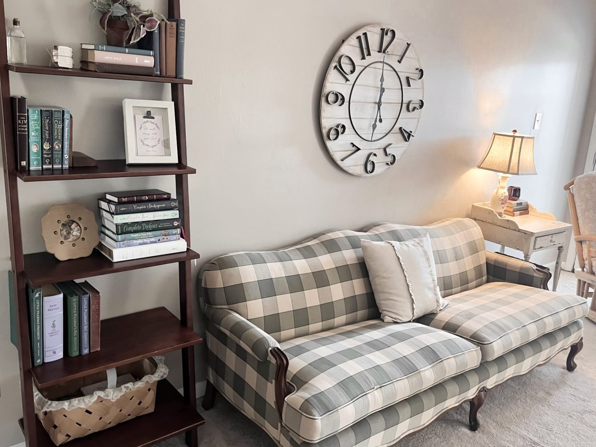 Country cottage living room, tall bookshelf with books and baskets, green checkered vintage couch with ruffle pillow large farmhouse clock
