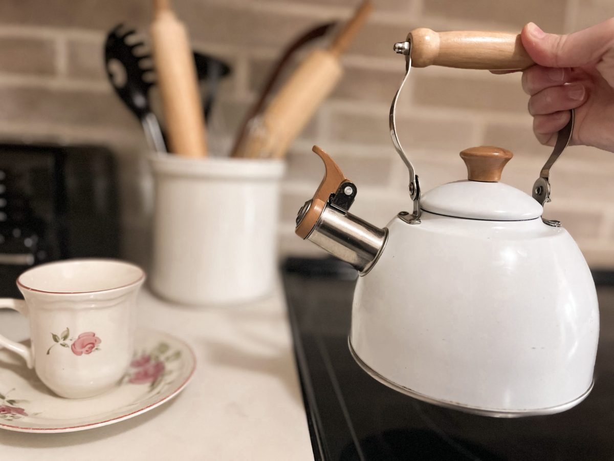 country cottage white tea kettle next to floral tea cup, crock of cooking utensils in background, grey brick black splash